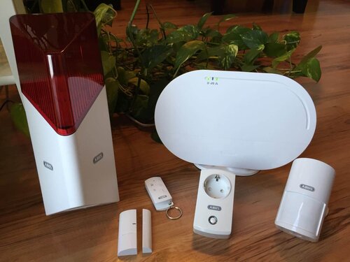 Das Bild zeigt ein modernes Alarmsystem mit Bewegungsmelder, Zentraleinheit und Sensoren auf einem Holztisch.