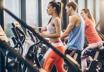 Das Bild zeigt ein Fitnessstudio von innen und 3 Menschen, die am Laufband stehen.